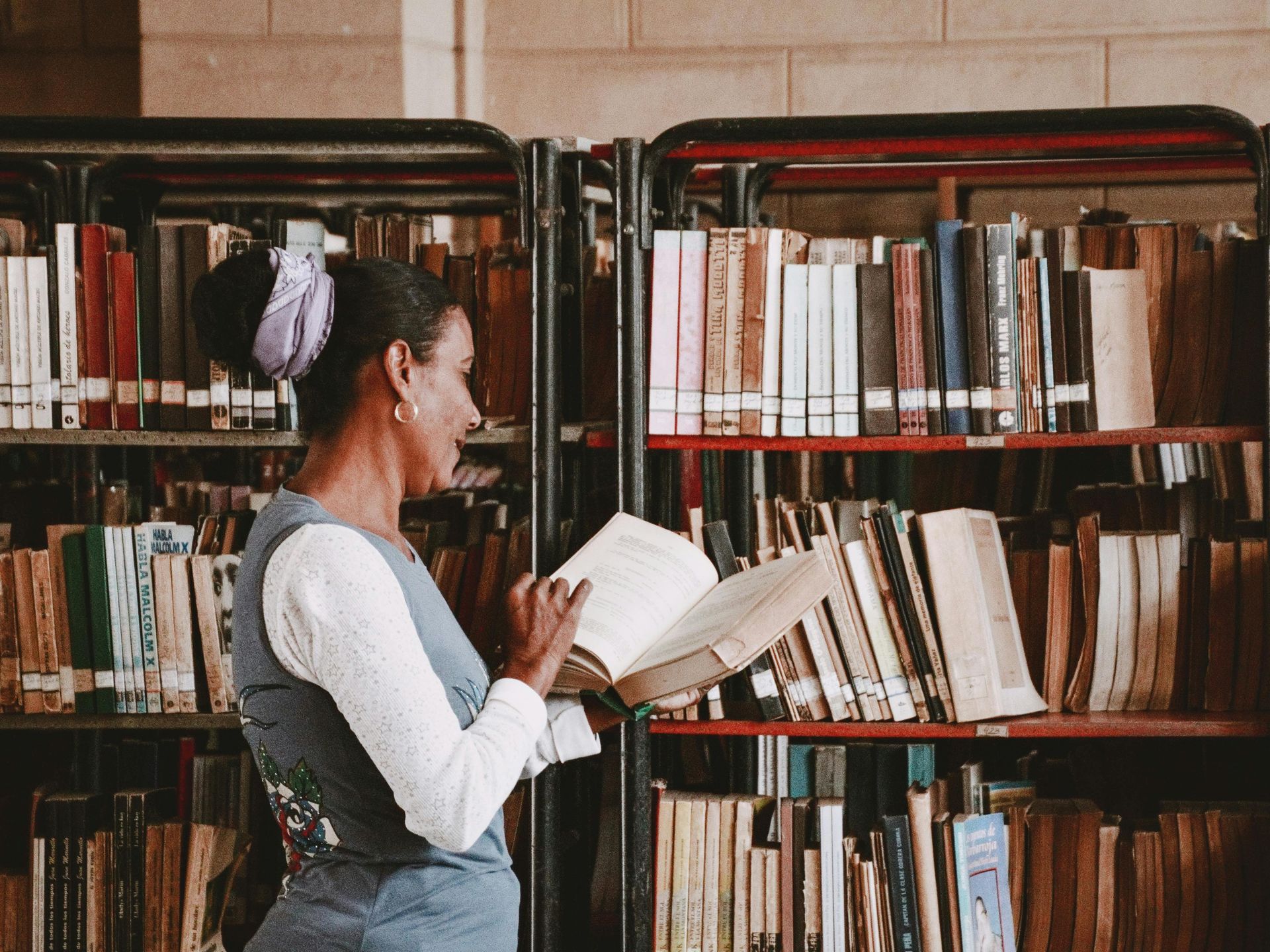 Woman reading an open book in a library aisle, with shelves of books behind her.