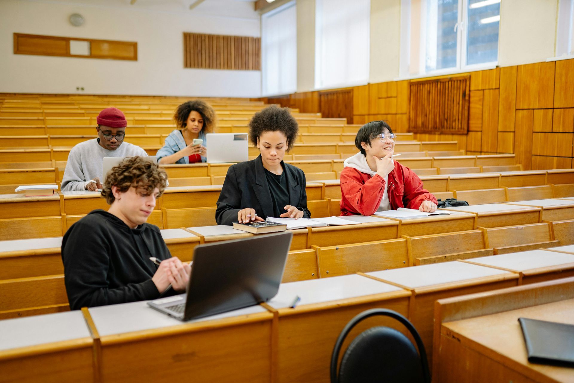 Students seated in a lecture hall, one using a laptop, others writing and listening attentively.