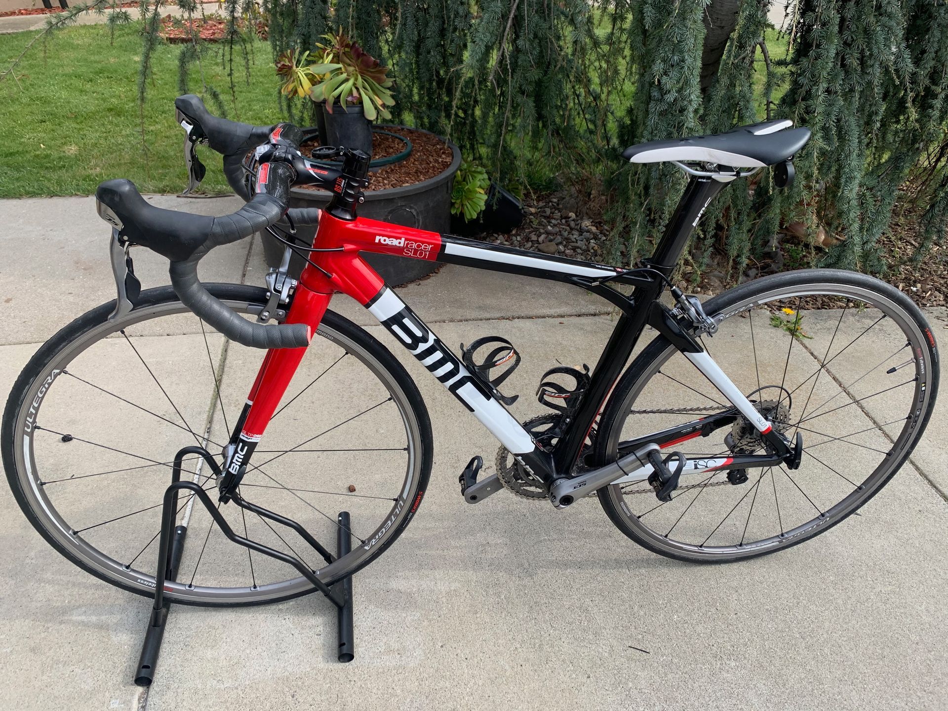 Red, white, and black BMC road bike on a bike stand, outdoors.