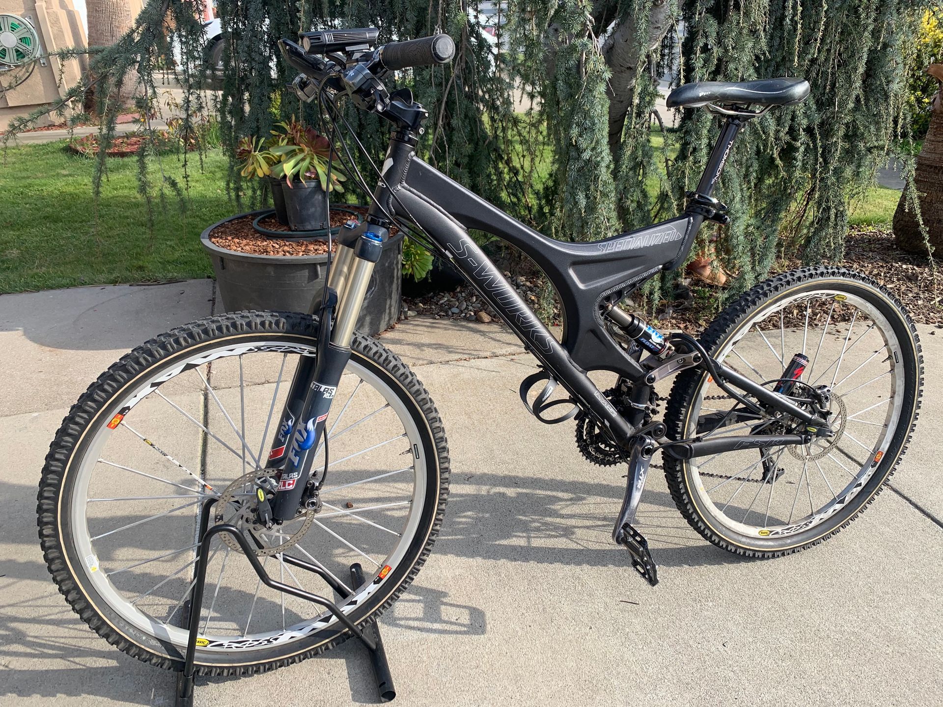 Black mountain bike on a stand outdoors, with white-rimmed wheels, suspension fork, and disc brakes.
