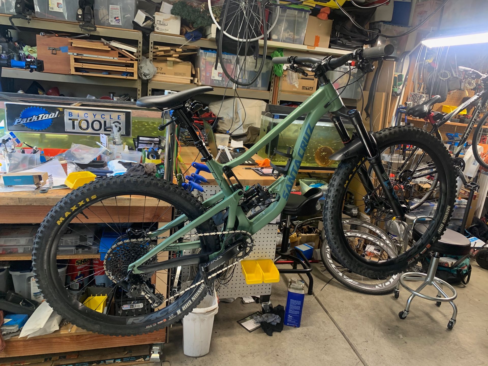 Green mountain bike in a cluttered garage, angled view, gear shift and suspension visible.