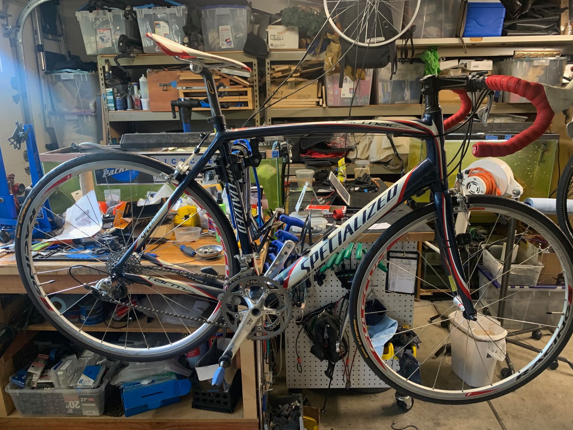 Specialized road bike in a cluttered workshop, black frame, red handlebar tape, silver wheels.