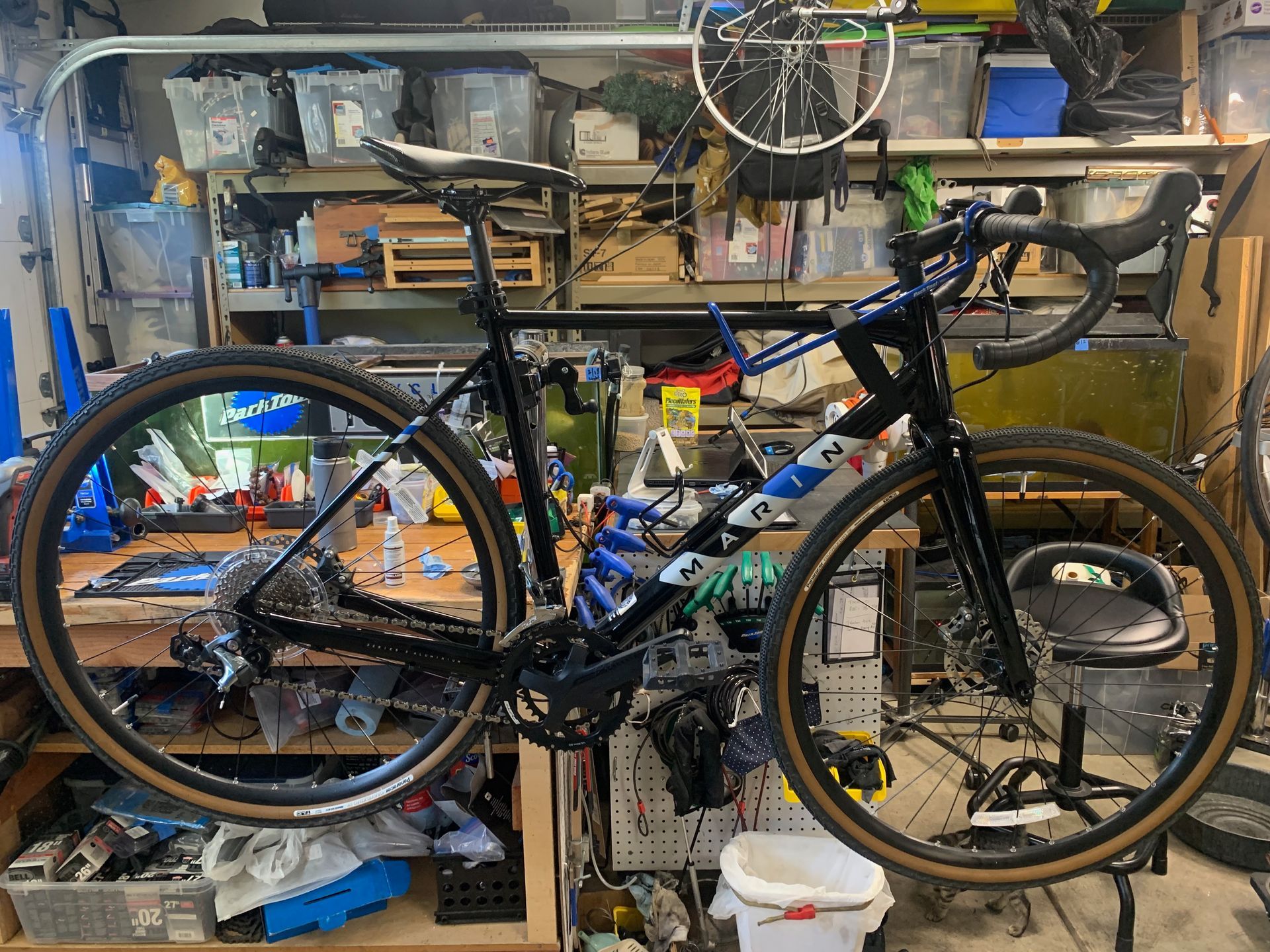 Black and blue gravel bike, tan tires, in a cluttered garage, bike repair tools in background.