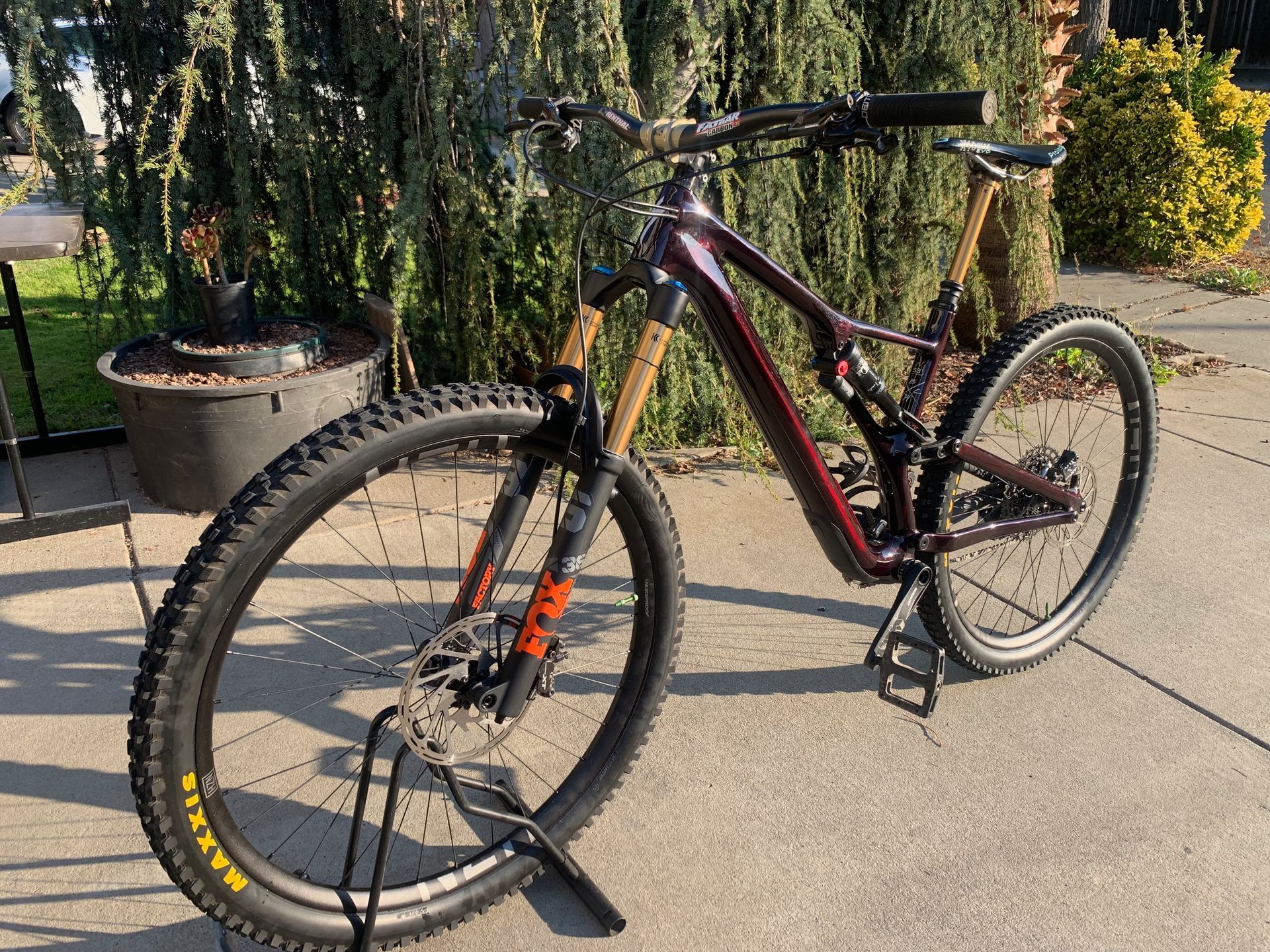 Dark red mountain bike parked on a paved surface with a green hedge backdrop.