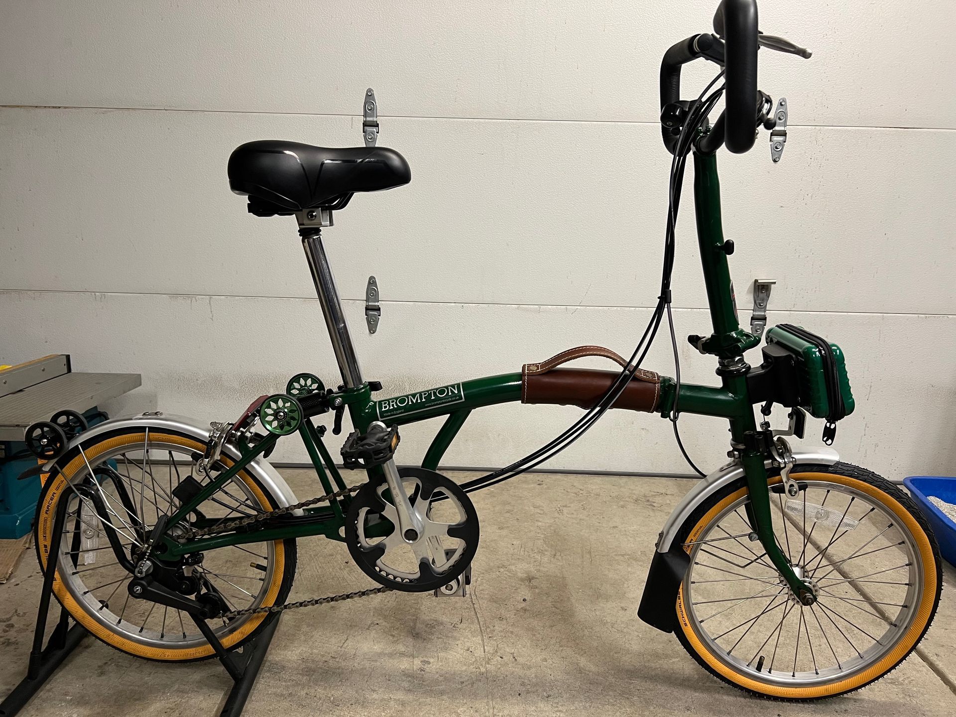 Green folding Brompton bicycle with tan tires on a black stand.
