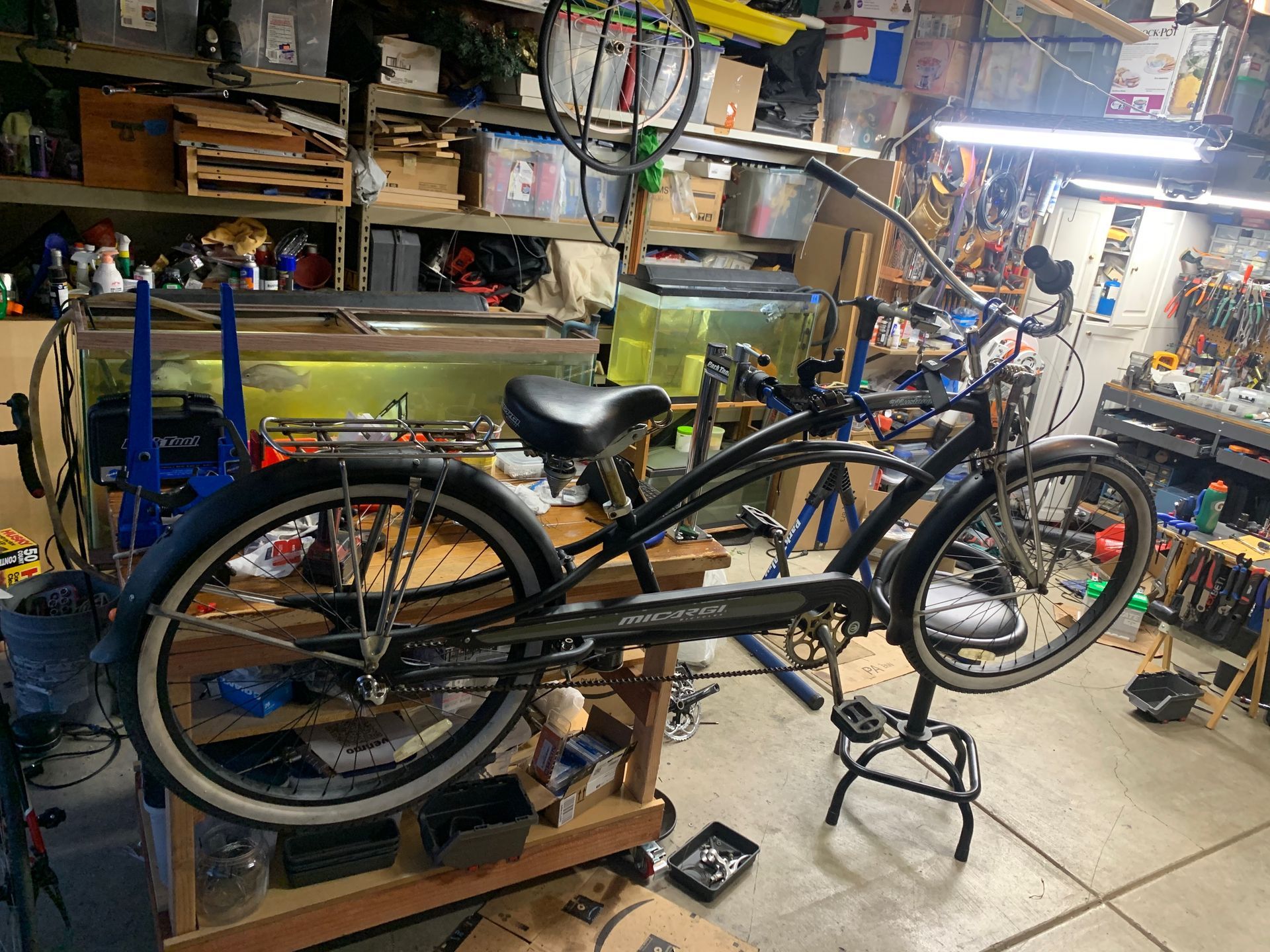 Black cruiser bicycle on a repair stand in a cluttered workshop. White-walled tires, black fenders.