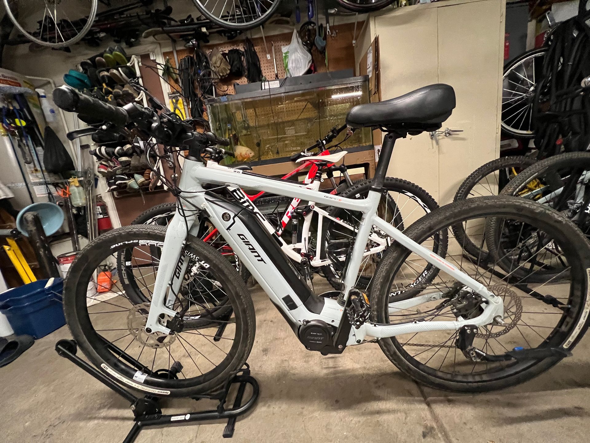 Light gray e-bike on a stand, in a cluttered garage, with other bikes visible in background.