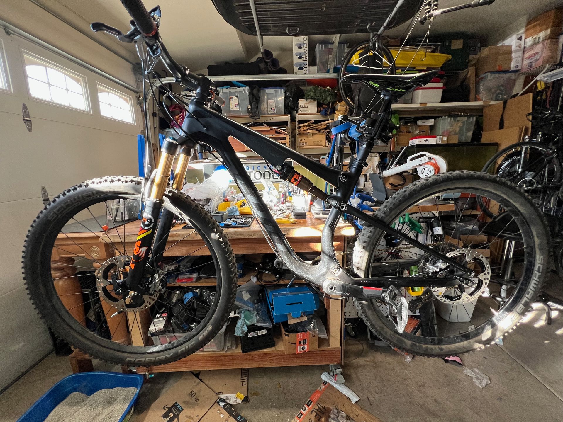 Mountain bike in a garage, black frame, gold suspension, wheels, tools, and a cluttered background.