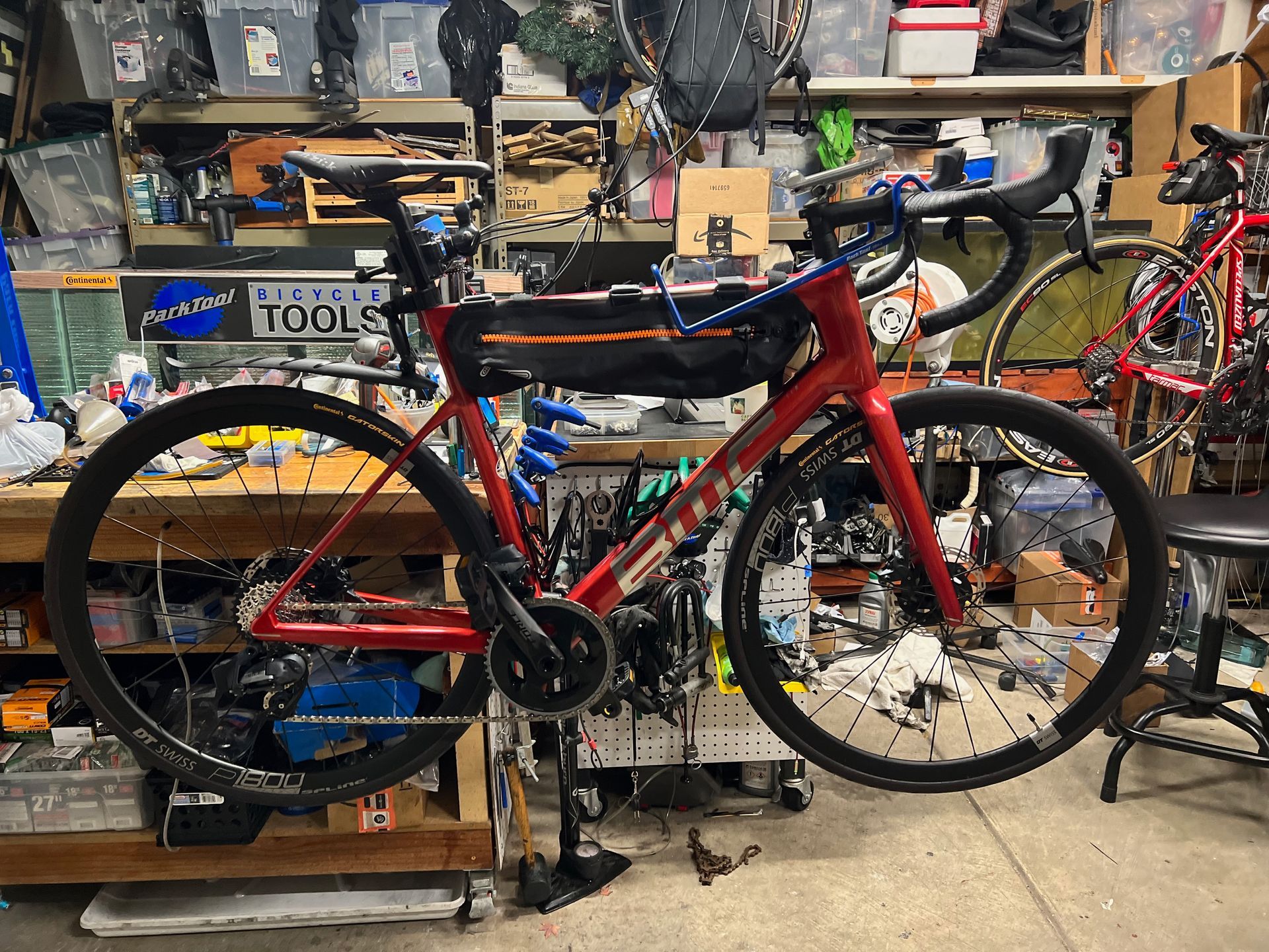 Red road bike in a workshop, black wheels, equipped with a top tube bag, and on a repair stand.