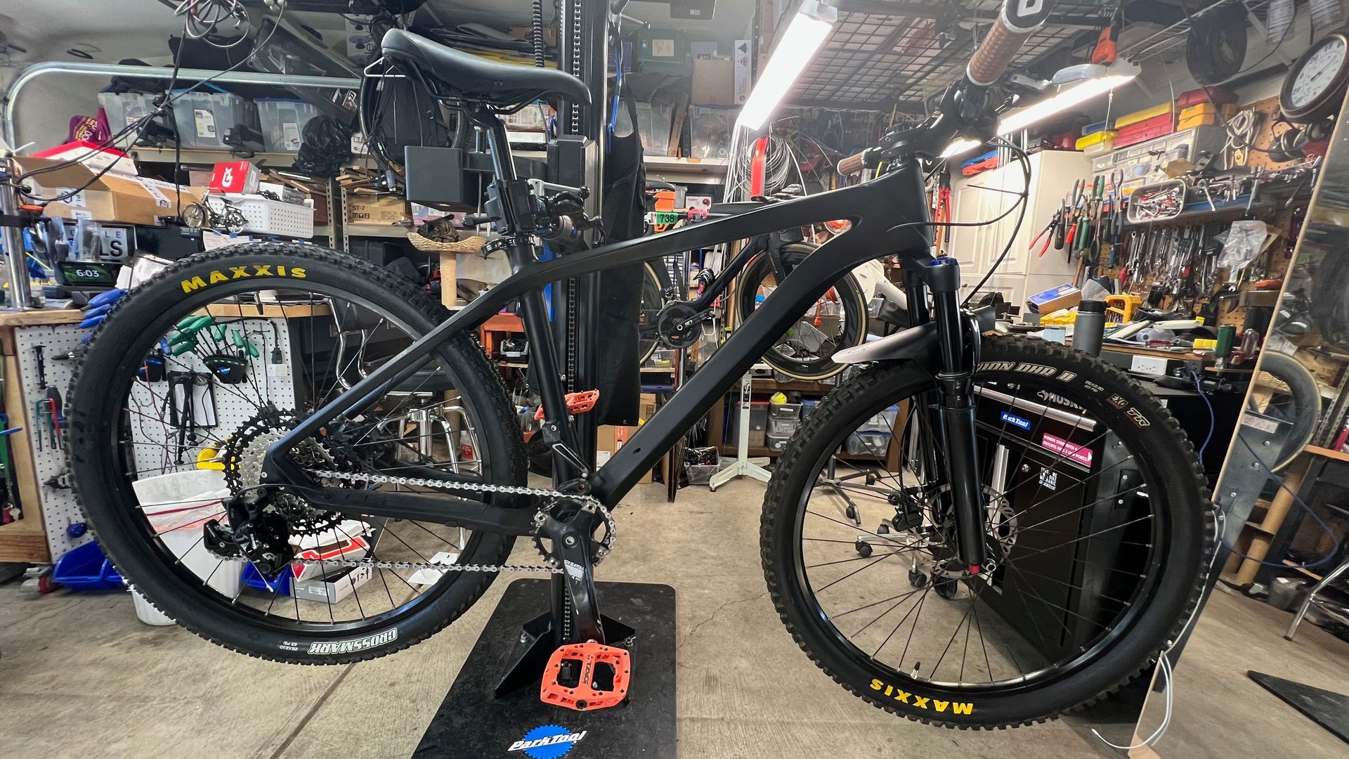 Black mountain bike on a stand with orange pedals, in a cluttered workshop.