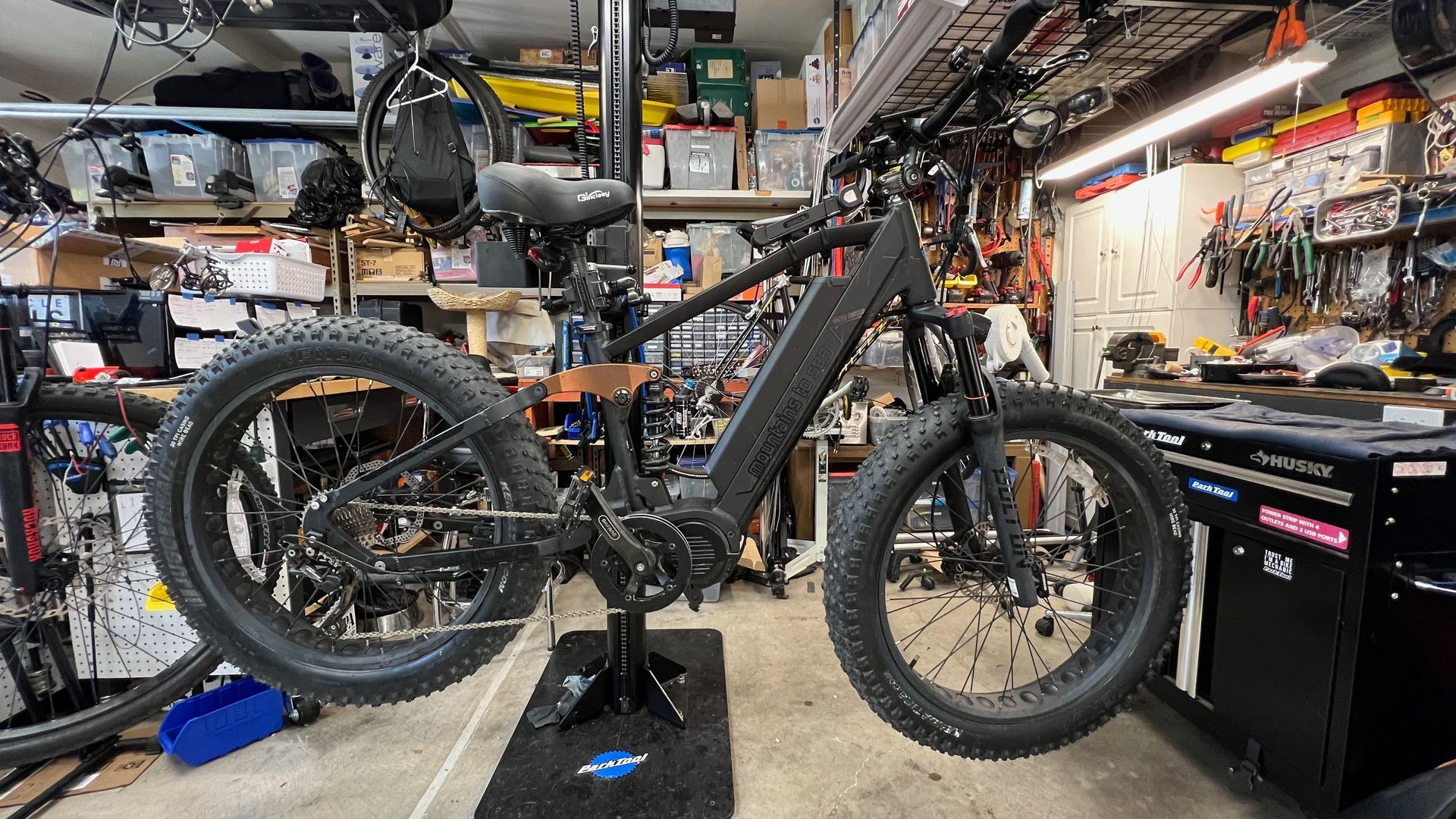 Black fat-tire e-bike on a repair stand in a cluttered garage with tools and parts.
