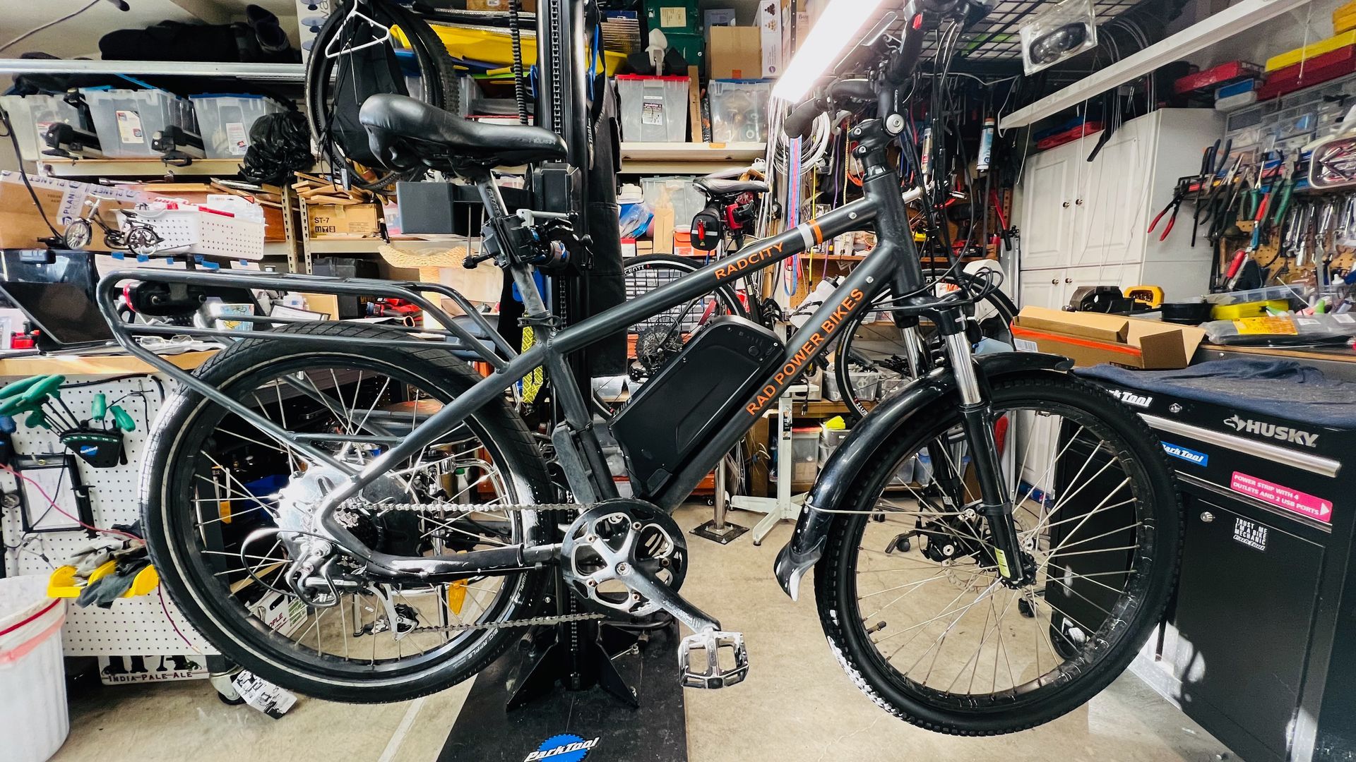 Electric bicycle in a workshop setting, gray frame, black fenders, and battery pack.
