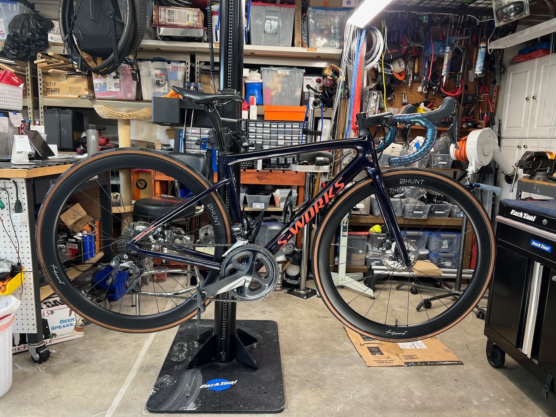 Road bike on a repair stand in a cluttered garage, with blue handlebars and tan tires.