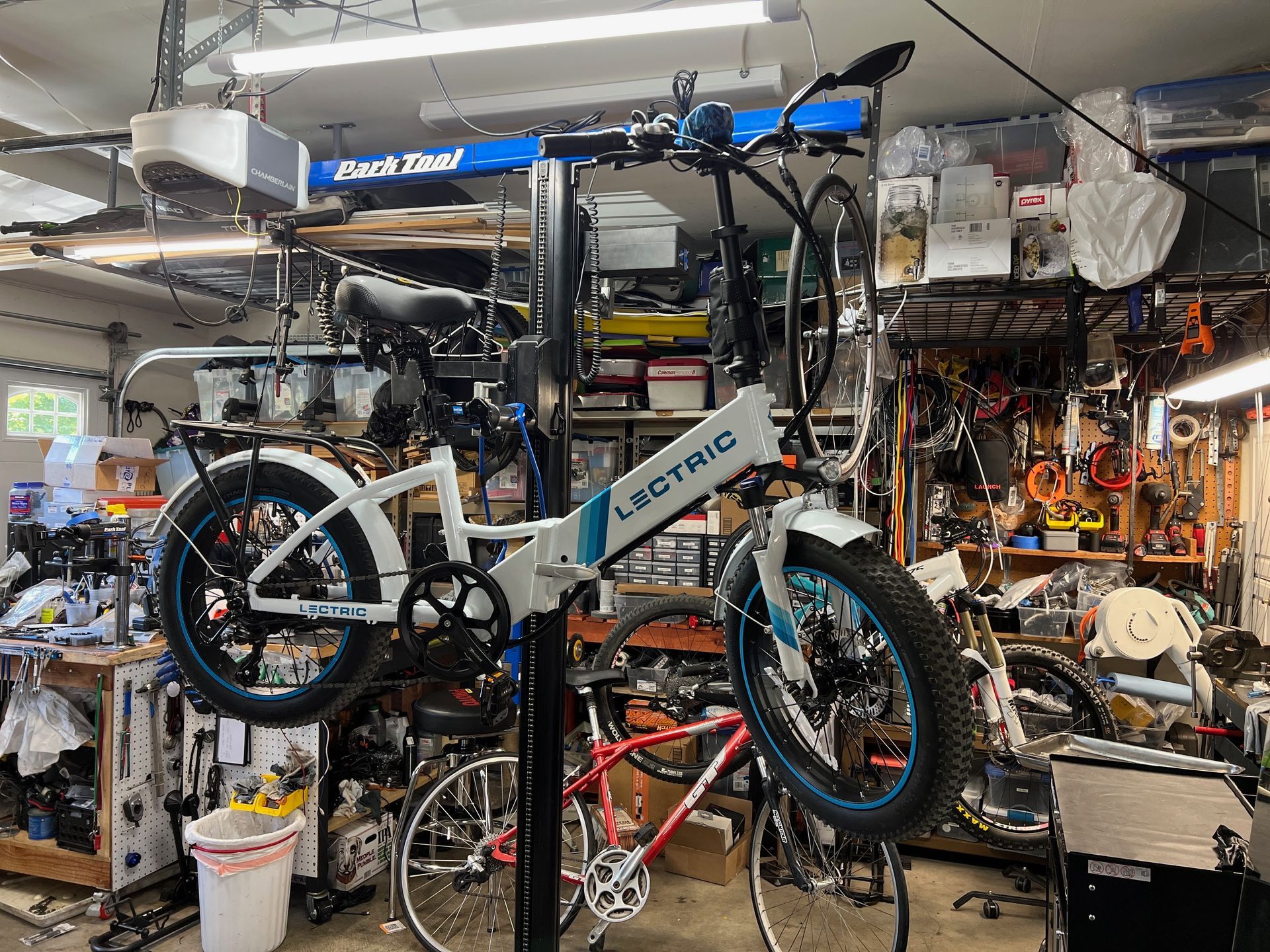 White and blue Lectric e-bike on a repair stand in a cluttered garage, other bikes visible.