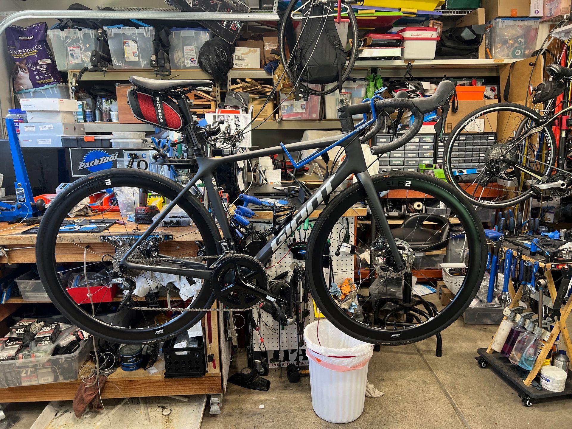 Black and gray bicycle hanging in a workshop; cluttered shelves in the background.