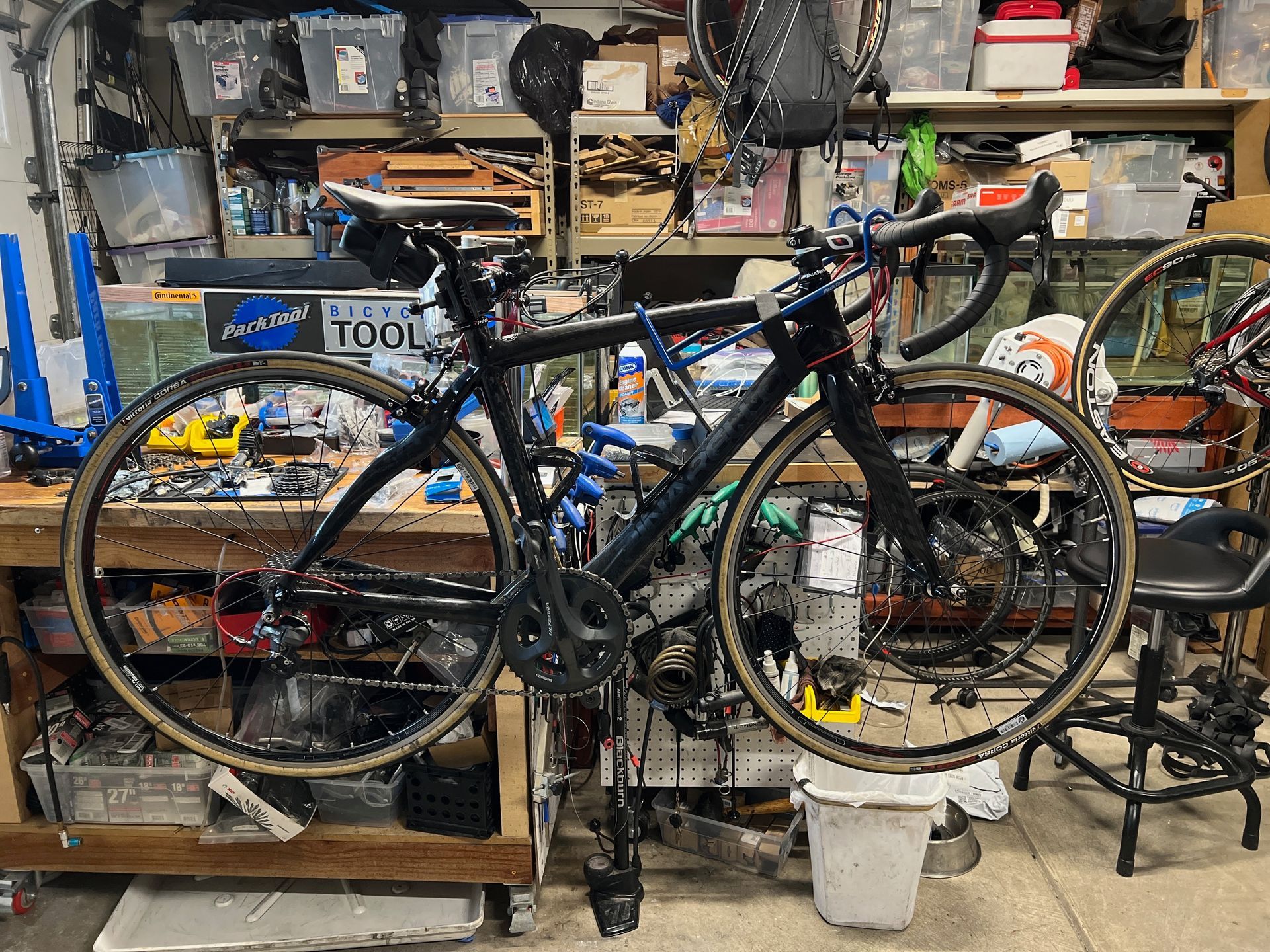 Black road bicycle in a cluttered workshop, wheels off the ground, tools and parts visible.