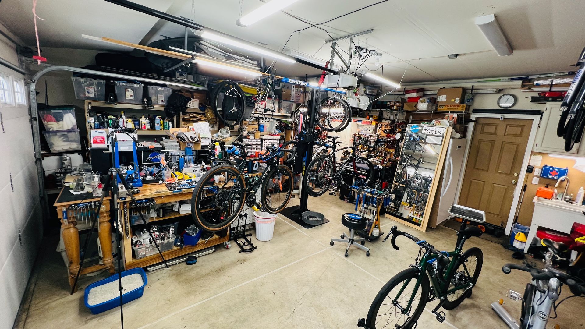 Cluttered garage with many bicycles, tools, and equipment, set up as a repair and maintenance workspace.
