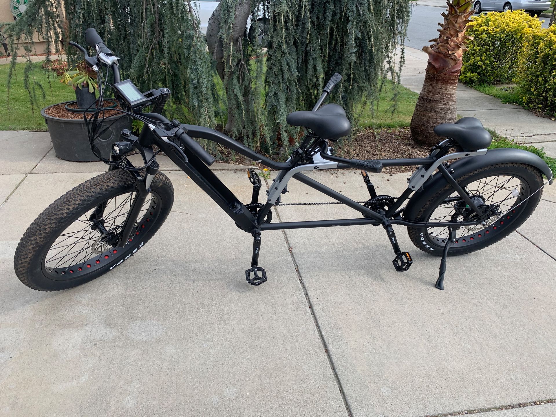 Black tandem electric bike on a sidewalk, two seats, fat tires.