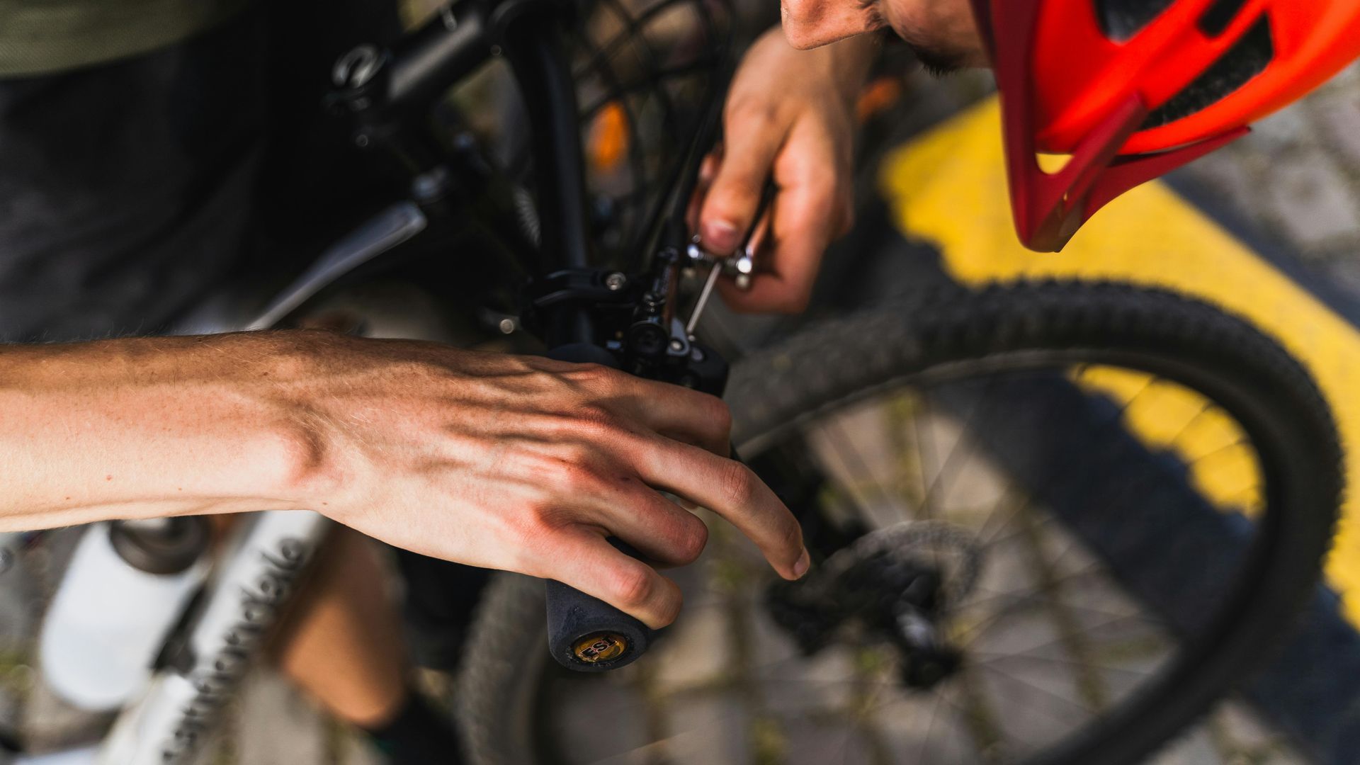 A person wearing a red helmet uses a tool to adjust the handlebars of a Cannondale bicycle on a cobblestone surface.