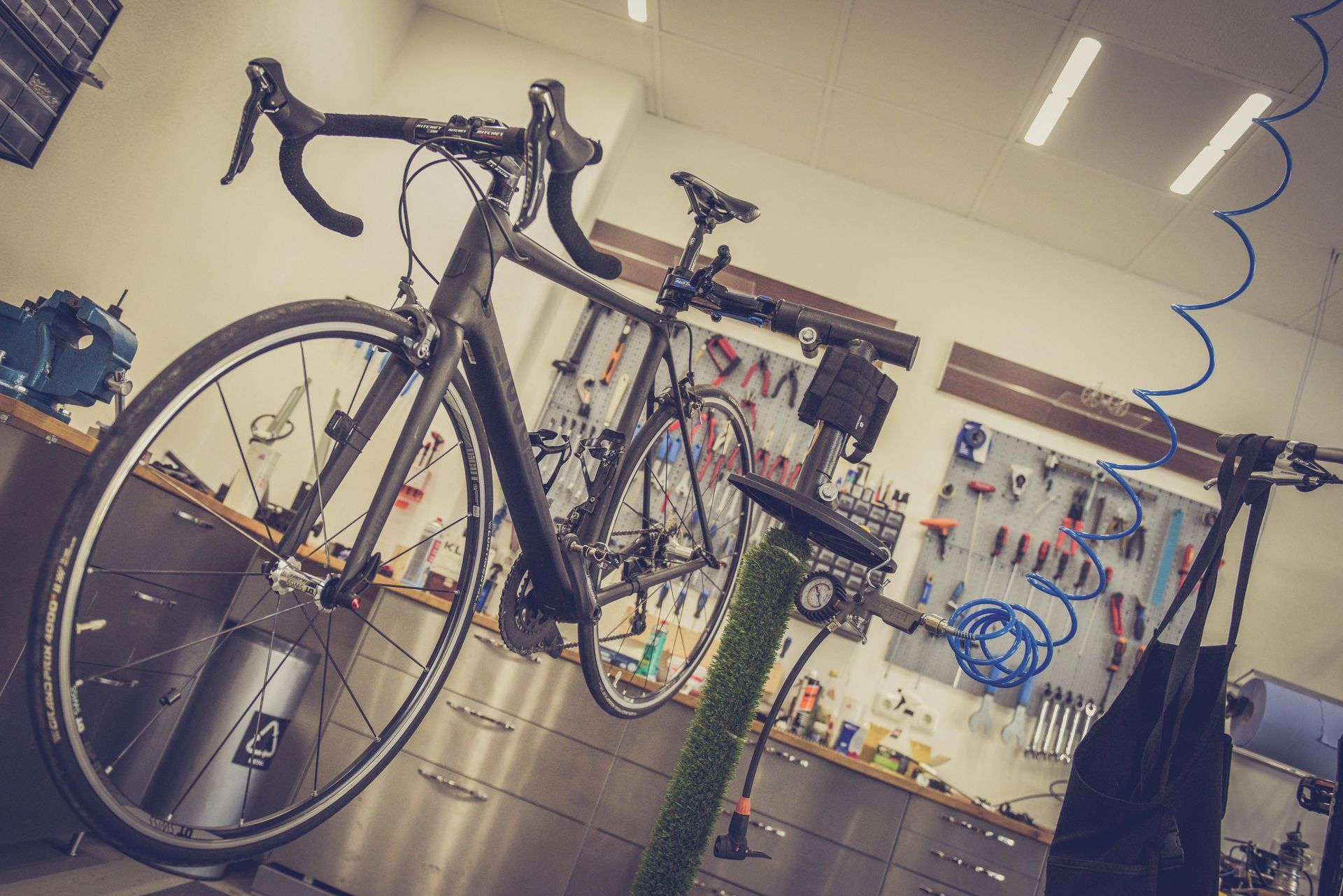 A black road bike mounted on a stand inside a bicycle repair shop, with tools organized on a wall in the background.