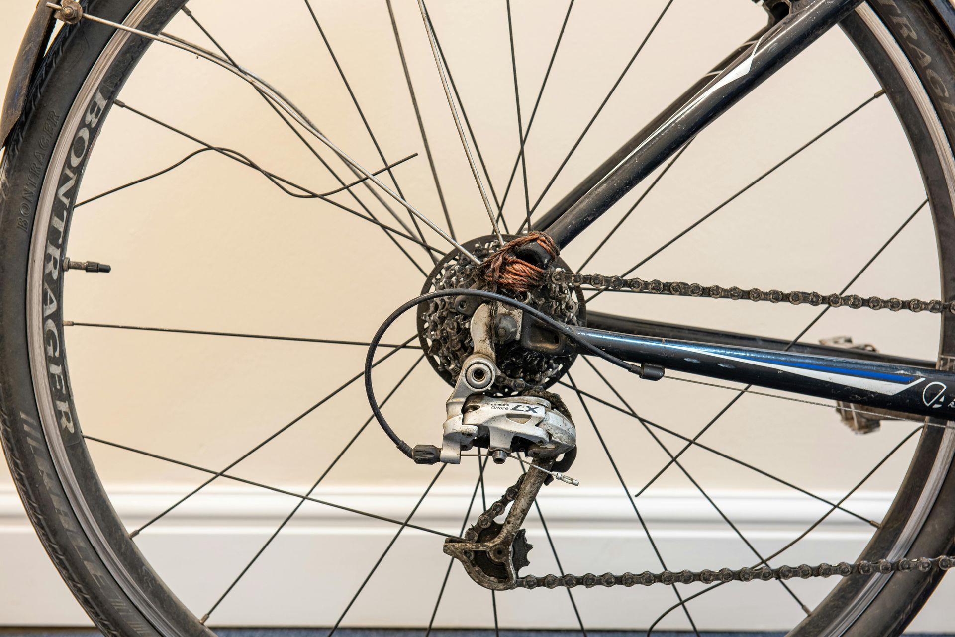 Close-up of a bicycle’s rear wheel, showing the gears, rear derailleur, chain, and metal spokes against a light wall.