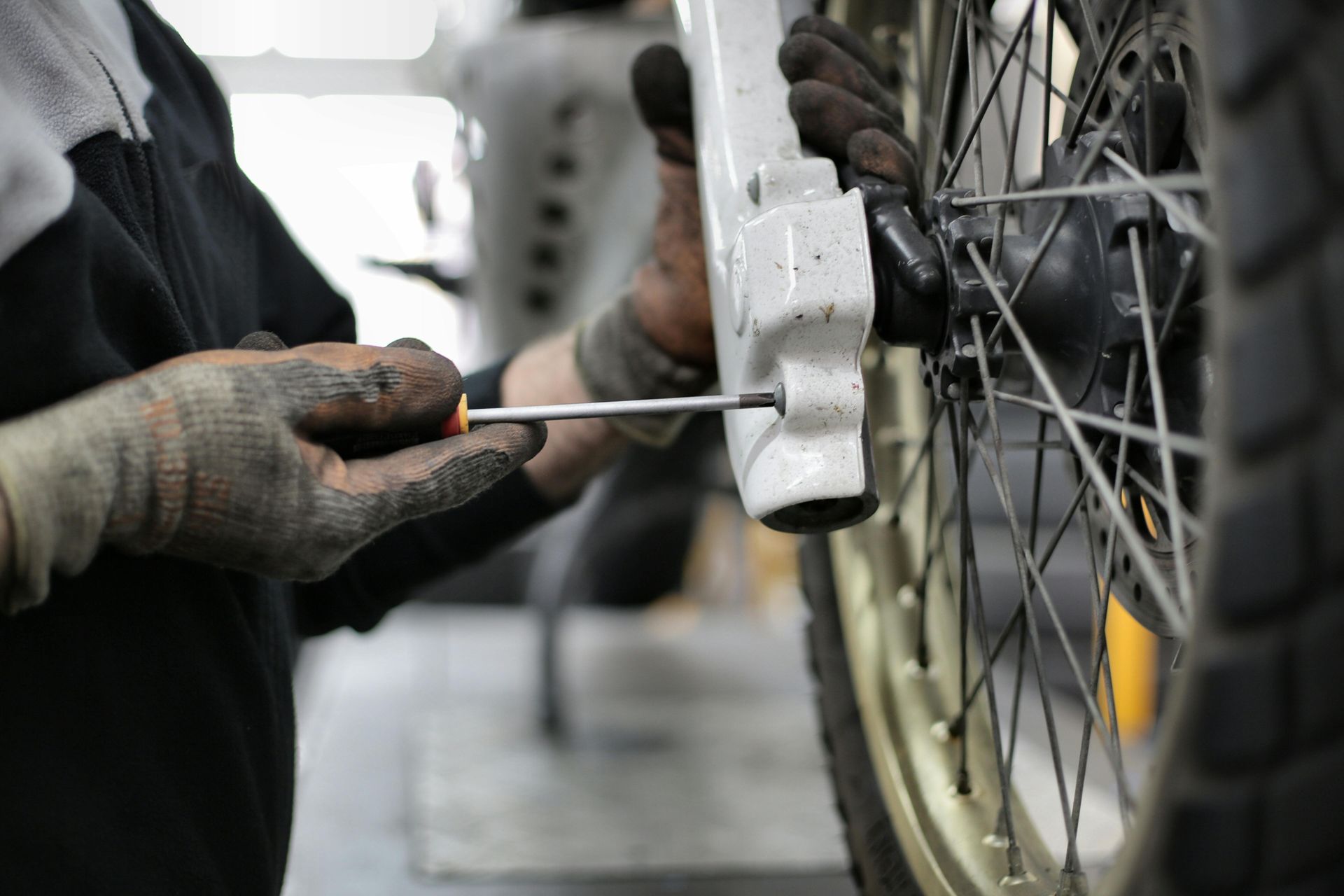 A mechanic wearing soiled gloves uses a screwdriver on the front fork of a motorcycle wheel in a workshop.