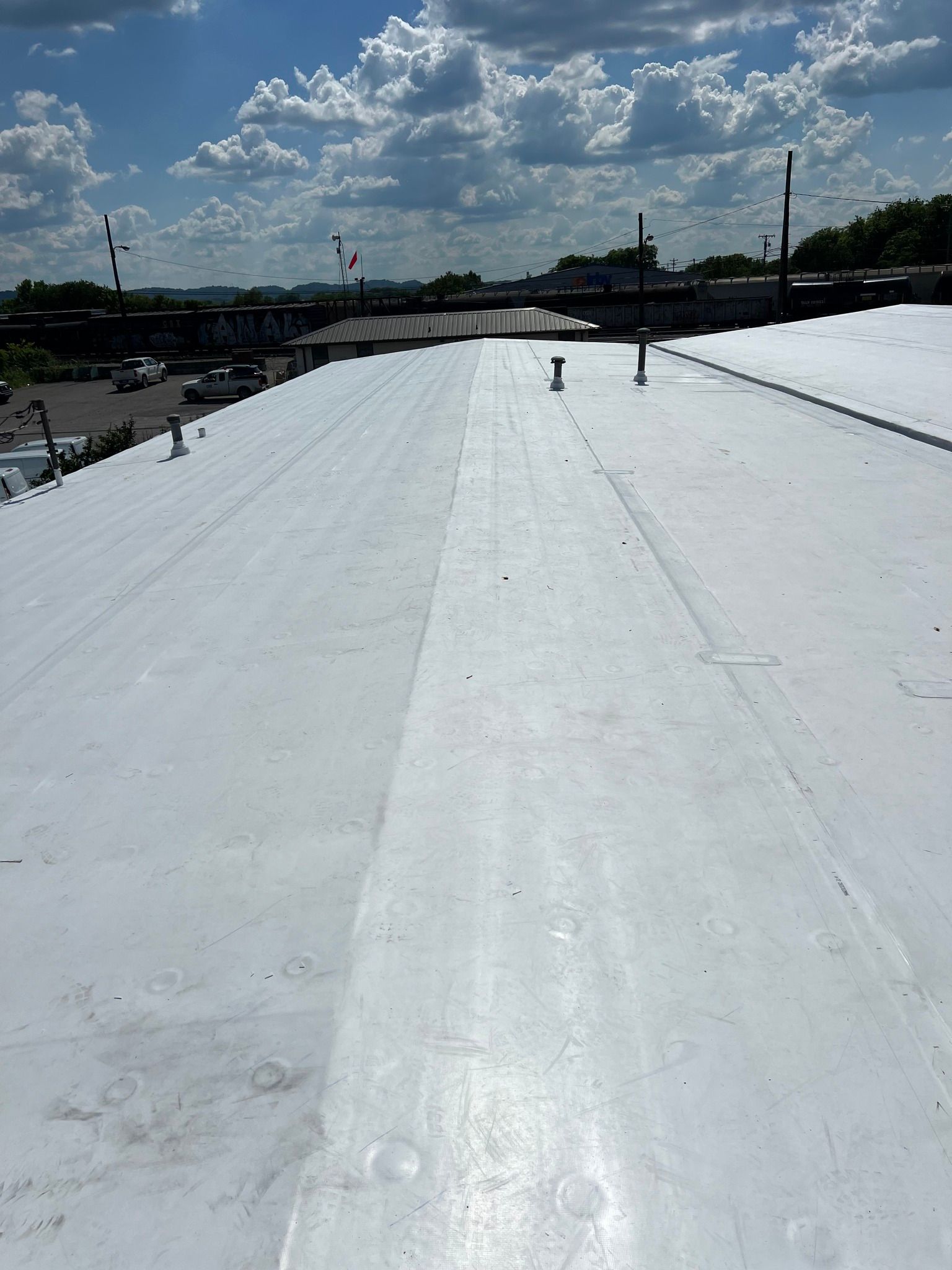 White commercial roof with seams, pipes, and distant buildings under a partly cloudy sky.