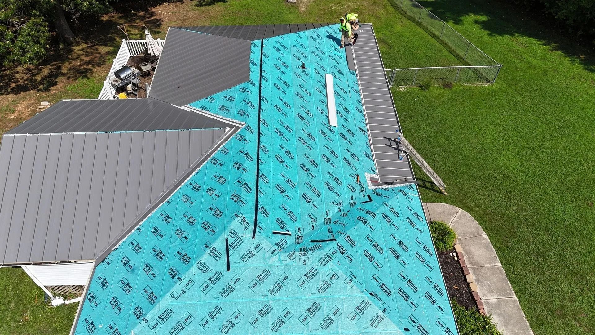 Overhead view of a house roof partially covered with turquoise underlayment, a worker installing it.