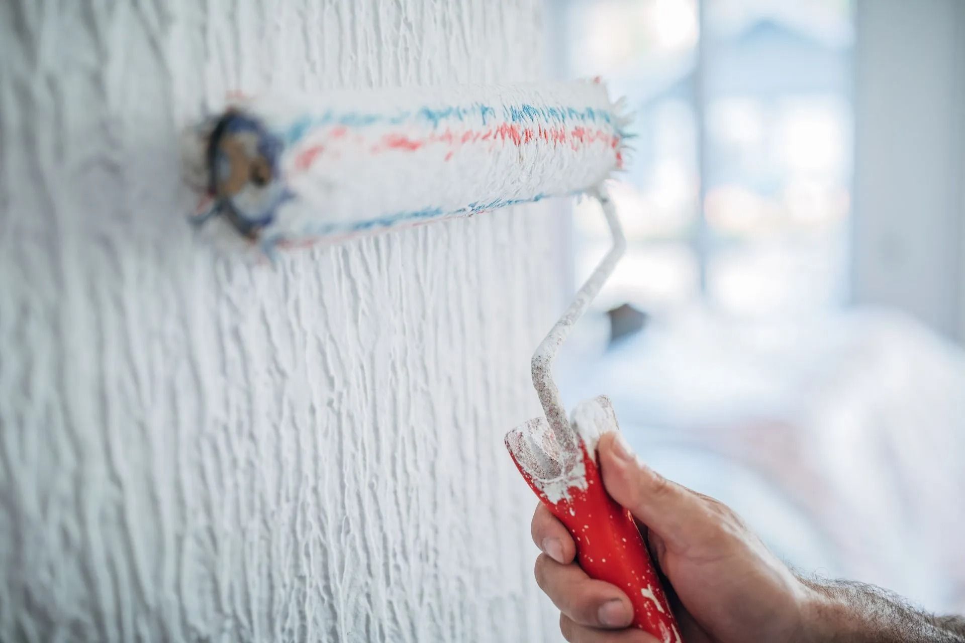 Hand holding paint roller applying white paint to a textured wall.