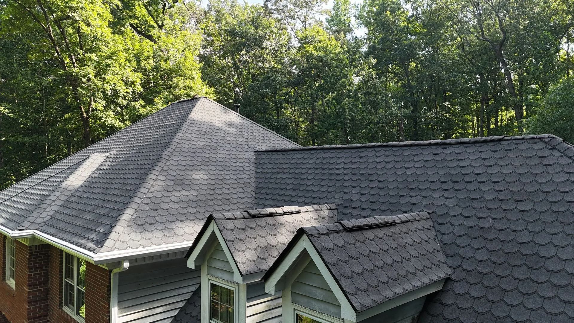 A gray shingled roof with a dormer and chimney, set against a backdrop of green trees.