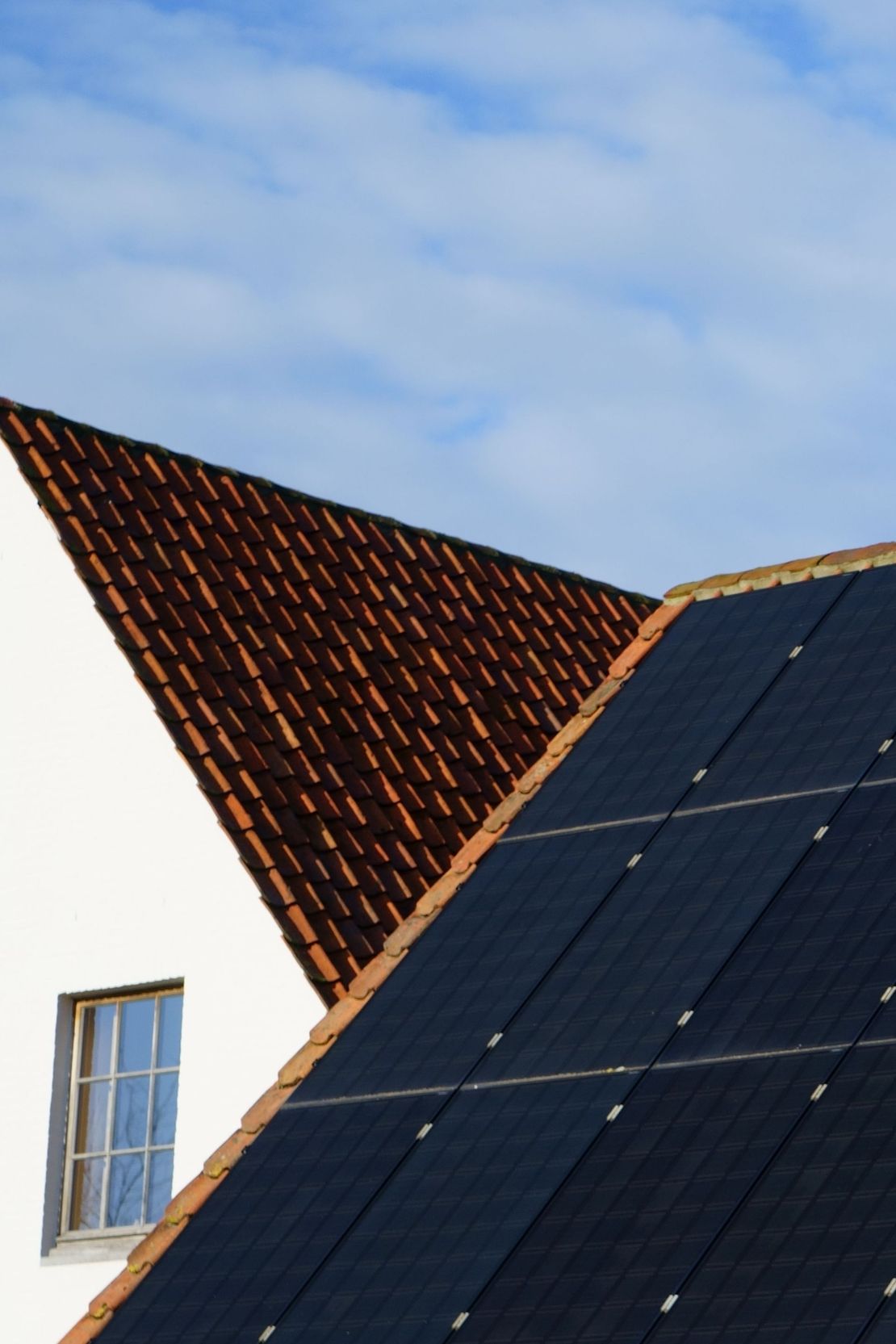 Solar panels on a rooftop beside a triangular red-tiled roof under a blue sky.