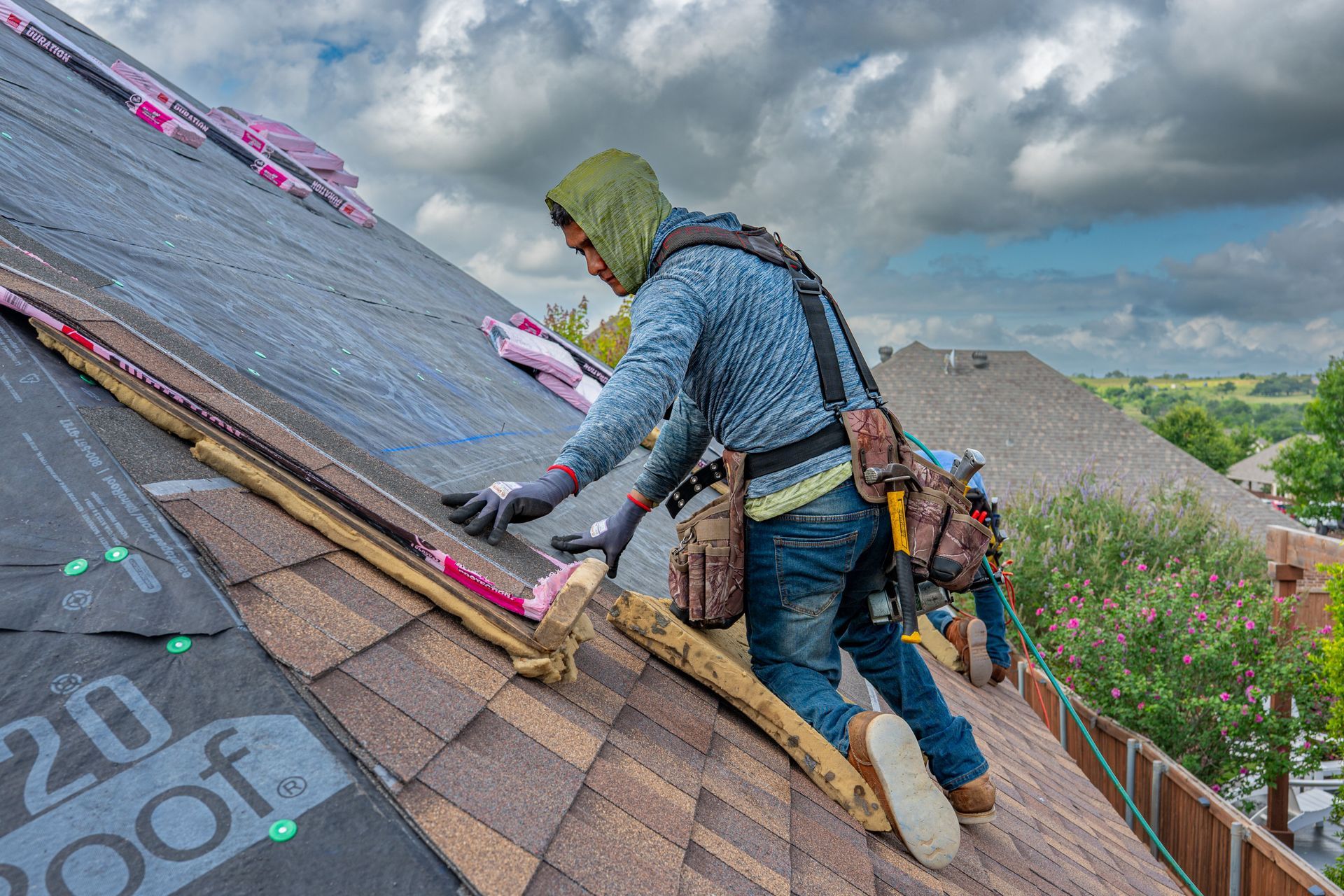 Worker repairing a shingled roof on a cloudy day, carrying tools and kneeling on the slope.