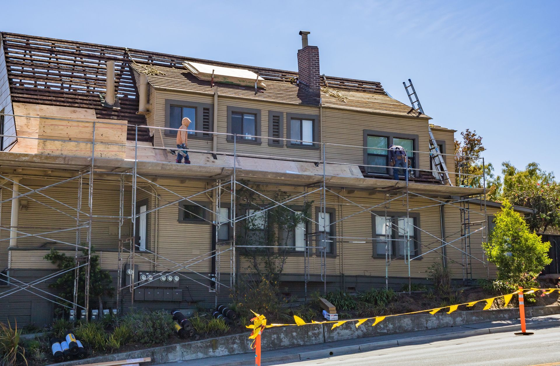 Two-story house under renovation with scaffolding, a ladder, and workers on the roof and balcony