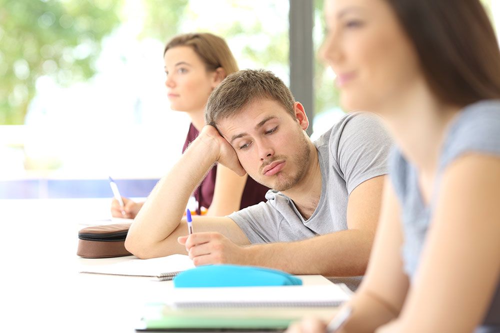Bored student in a classroom rests head on hand; two other students take notes at their desks.