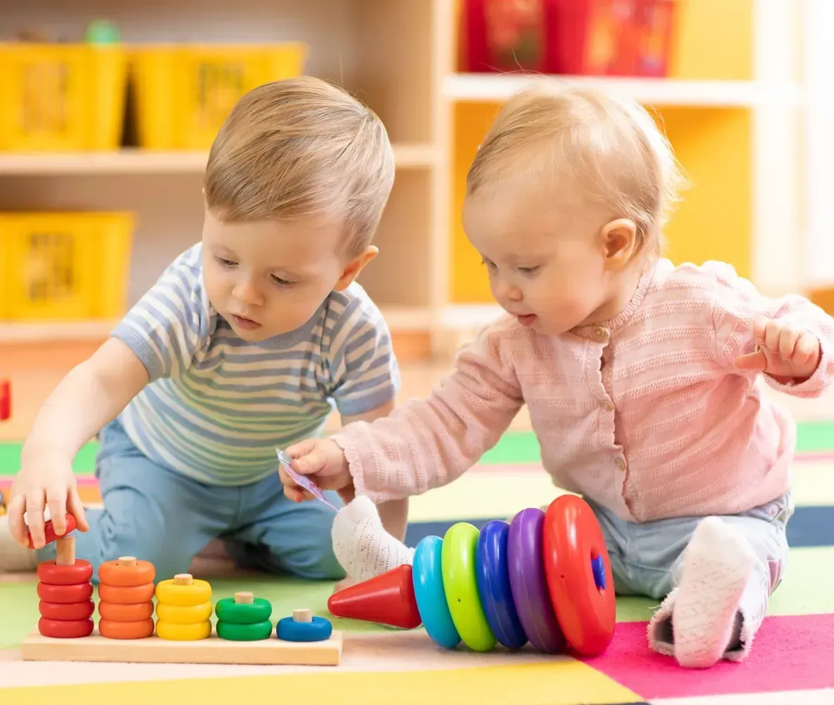 Two toddlers playing with colorful toys on a mat in a bright playroom
