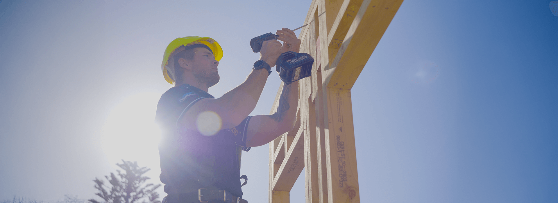 Construction worker using a drill on a wooden frame under a bright blue sky.
