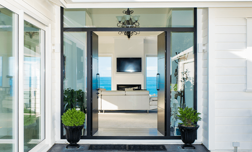 Glass front doors open to a living room with ocean view, flanked by plants.