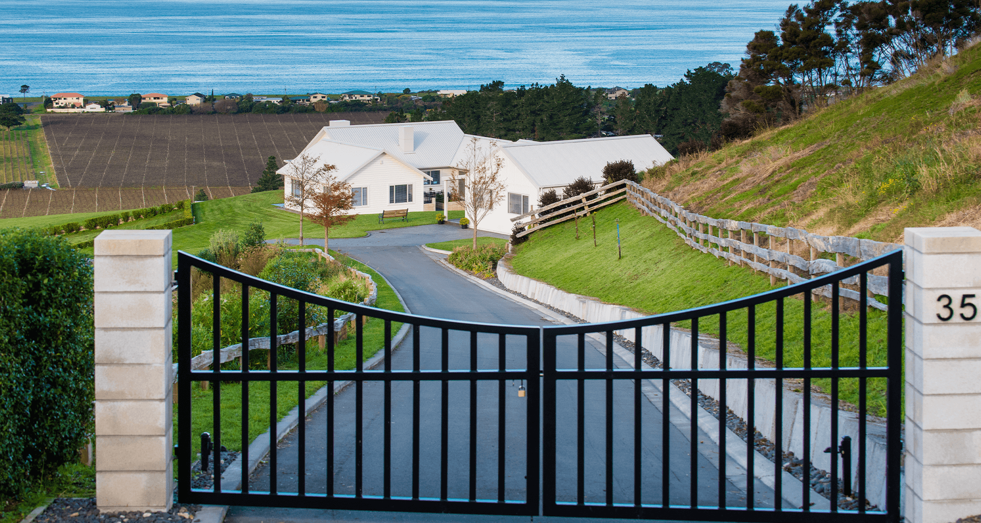 Black gated driveway leads to a white house on a hillside with ocean view.