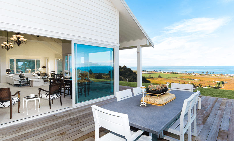 White house with open sliding doors leading to an outdoor dining area with ocean view.
