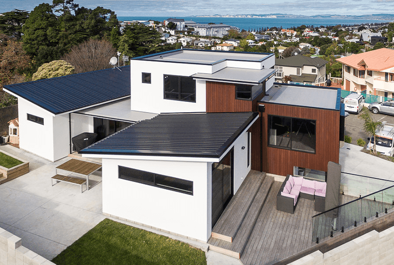 Modern white house with dark roof, wooden deck, and brown accent wall overlooking a city and water.