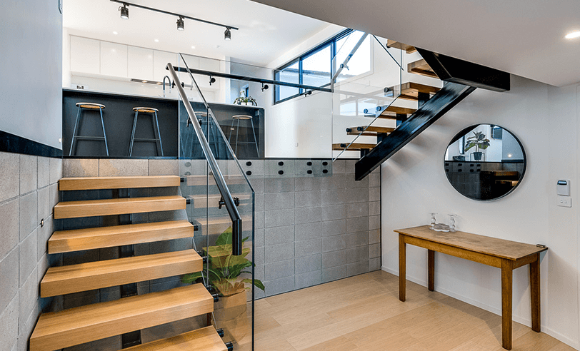 Interior view: wooden staircase leading to a kitchen with a black island and two stools, a round mirror on the wall.