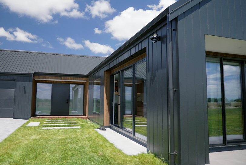 Modern single-story home with red textured exterior, chimney, and grassy lawn under blue sky.