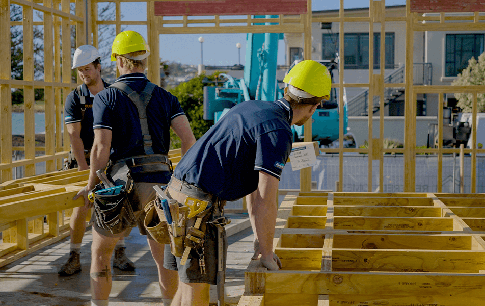 Construction workers assembling a wooden frame, wearing hard hats and work clothes in a sunny outdoor setting.