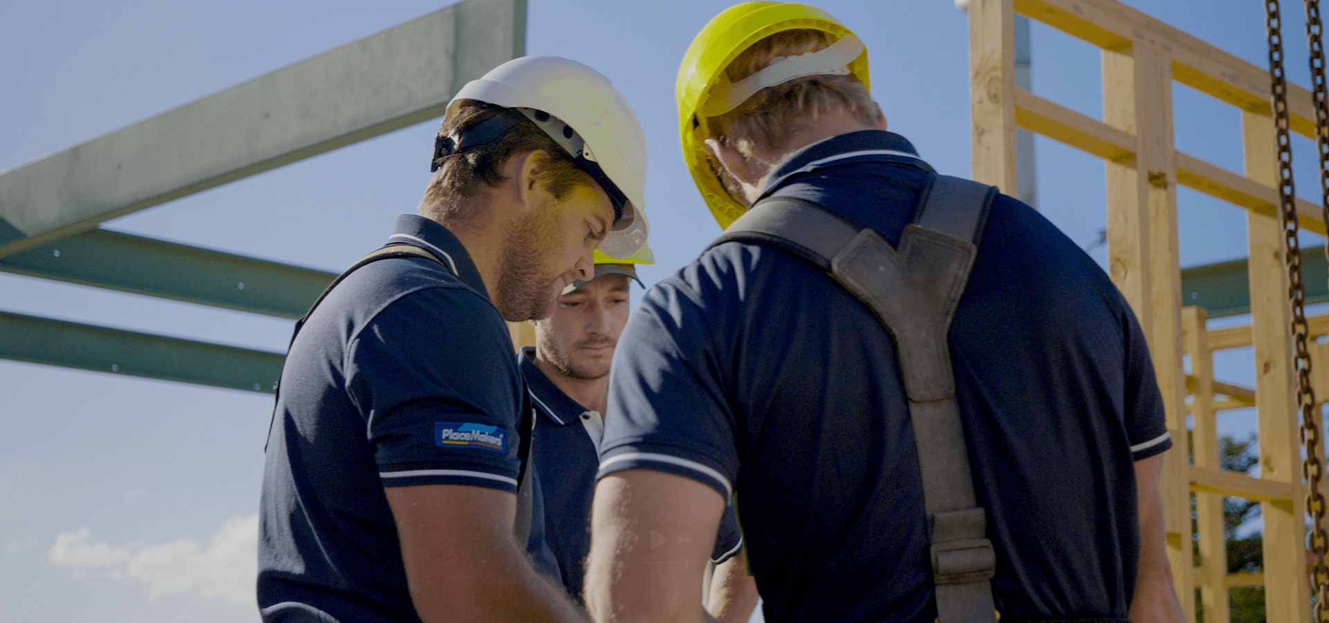 Three construction workers in hard hats discussing plans at a construction site, blue shirts.