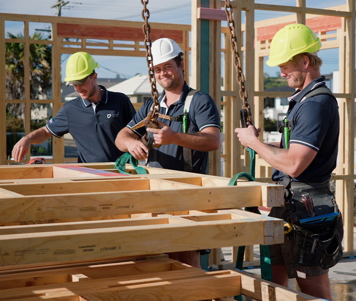 Three construction workers in safety gear lift wooden wall frame on a construction site.