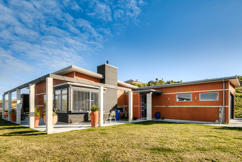Modern single-story home with red textured exterior, chimney, and grassy lawn under blue sky.