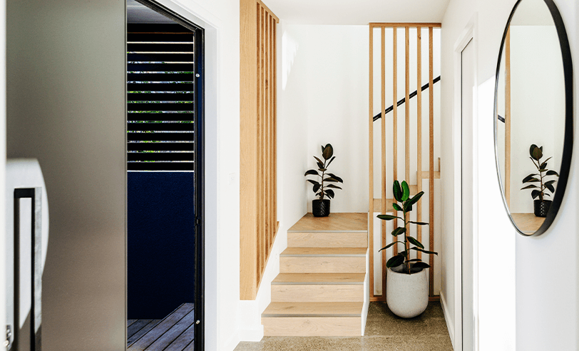 Entryway with wooden stairs, vertical wood slats, white walls, and a round mirror. Two potted plants.