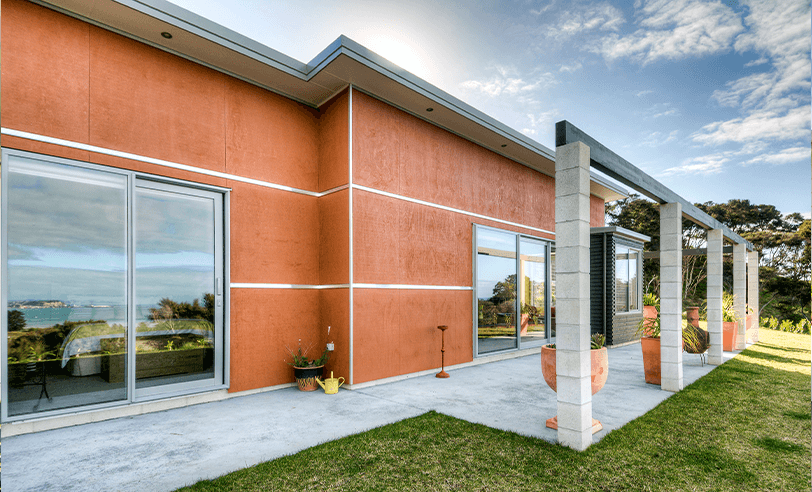 Terracotta-colored modern house with large windows, a patio, and a grassy lawn under a sunny sky.