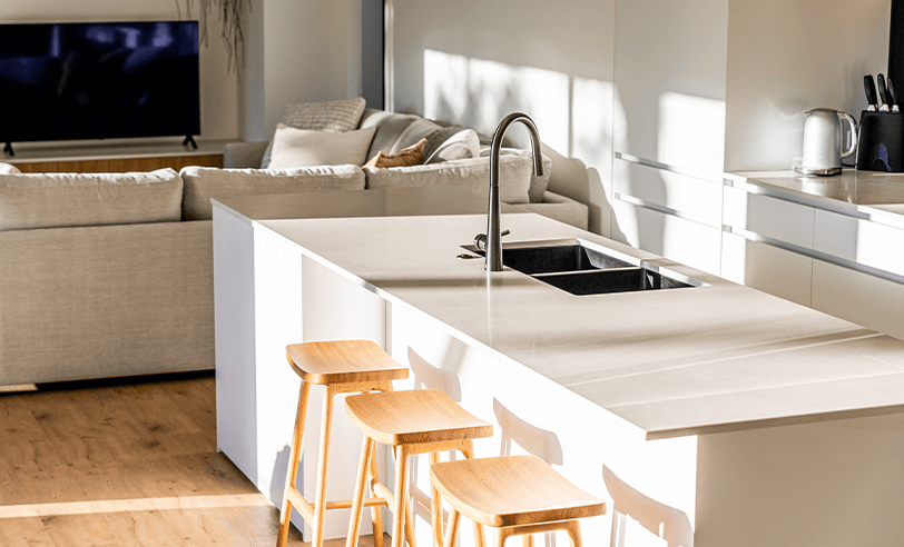 Modern kitchen with island, stools, and sink, overlooking a living room with a sofa and TV.