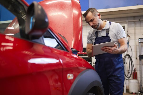 Mechanic in blue overalls examines a red car with its hood open, using a tablet in a garage. | King's Auto Center